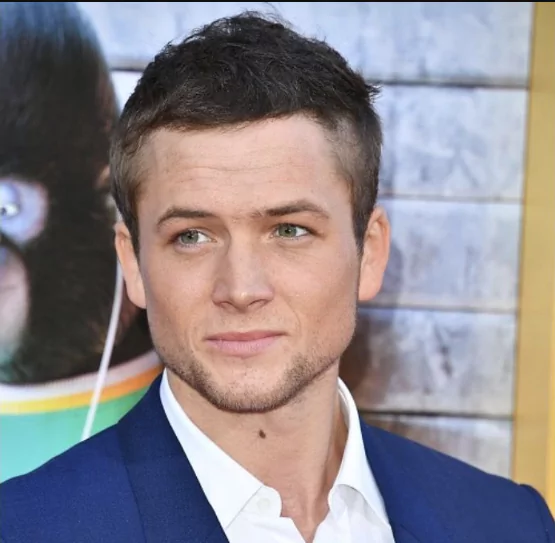 Taron Egerton: Medium Length Hairstyle With Widow's PeakTaron Egerton with a medium-length, swept-back hairstyle that accentuates his widow’s peak, styled naturally with soft volume and texture.Ask ChatGPT